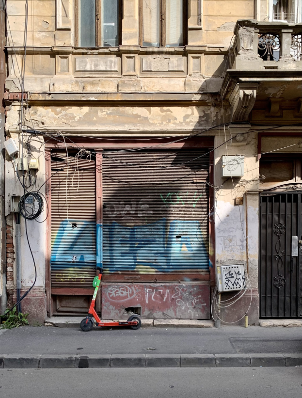Ein rotes Roller-Fahrzeug, das auf einer Straße neben einem Gebäude mit Fenstern, Läden und einem Tor geparkt ist, mit sichtbaren Kabeln und Gegenständen entlang der Straße und Text am unteren Bildrand.