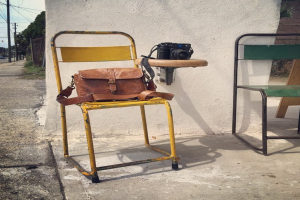 Eine Straße mit einer Wand davor, zwei Stühle auf der linken Seite mit einer braunen Tasche auf einem und einer Kamera auf dem anderen, sowie Strommasten, eine Straße und Bäume im Hintergrund.