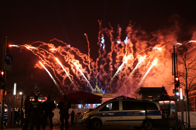 Polizeiauto vor Menschenmenge bei Silvesterfeierlichkeiten in Berlin geparkt, mit farbigen Feuerwerken am Himmel.