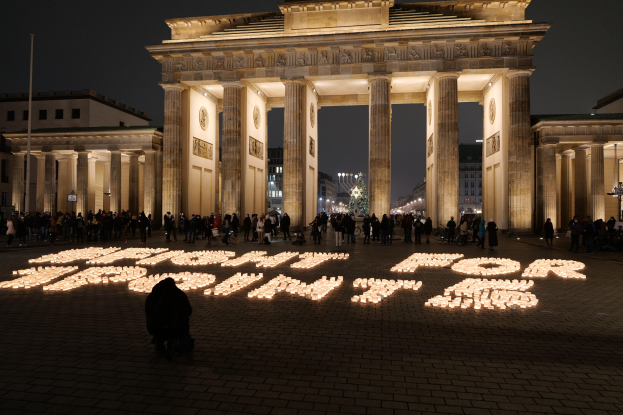 A group of people stands in front of the illuminated Brandenburg Gate in Berlin, Germany, with buildings, poles, and lights surrounding the area, and the words "Fight for Freedom" written on the ground.