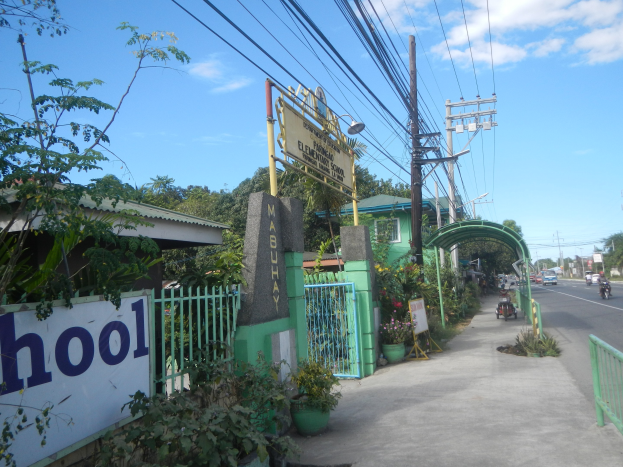 Einfahrt zu einer Schule mit einem "Schule"-Schild, Fahrzeuge auf der Straße, ein Tor, ein Eingang, ein Namensschild, Pflanzen, Bäume, Strommasten mit Drähten und eine bewölkte Himmel im Hintergrund.