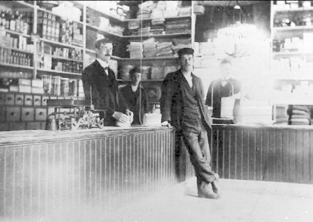 Black and white photo of three men at a store counter during what appears to be a robbery, with books and items in the background.