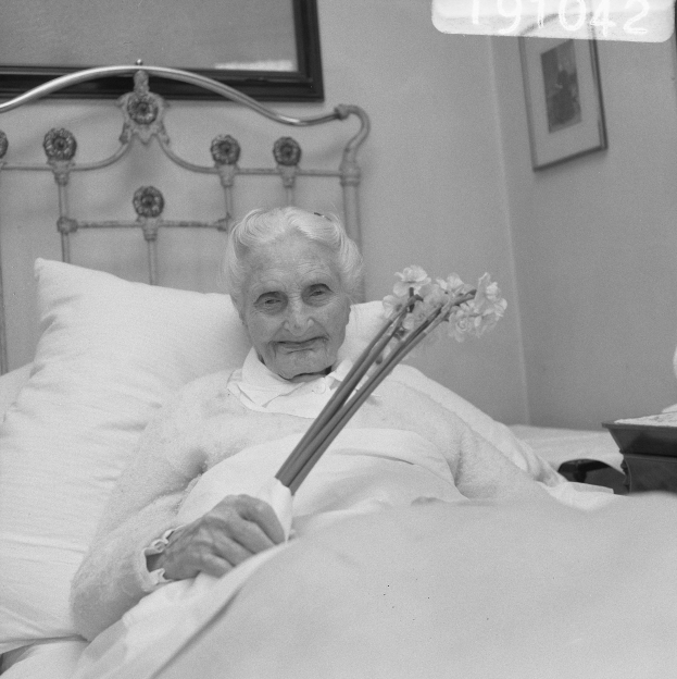 Alte Frau im Krankenhausbett liegend, die einen Gehstock hält, umgeben von Kissen, mit einem Tisch und Gegenständen rechts und gerahmten Bildern an der Wand im Hintergrund.
