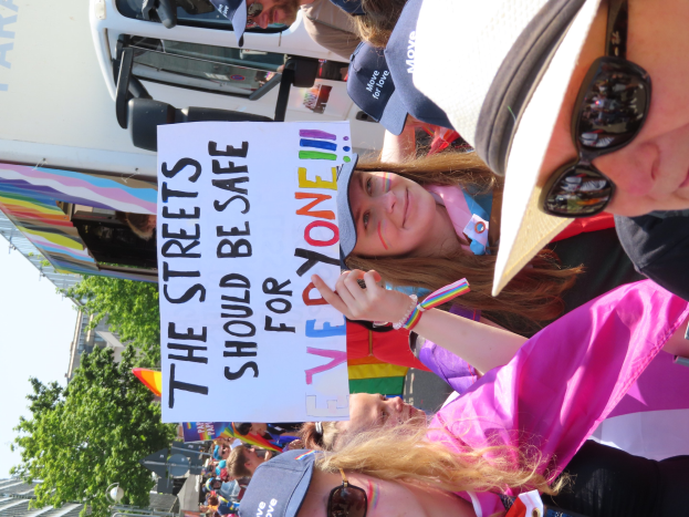 Gruppe von Menschen vor einem Lastwagen mit Schildern, darunter eines mit der Aufschrift "Die Straßen sollten sicher sein", einige tragen Mützen und Schutzbrillen, vor einem Hintergrund aus Bäumen, Gebäuden und einem klaren blauen Himmel.