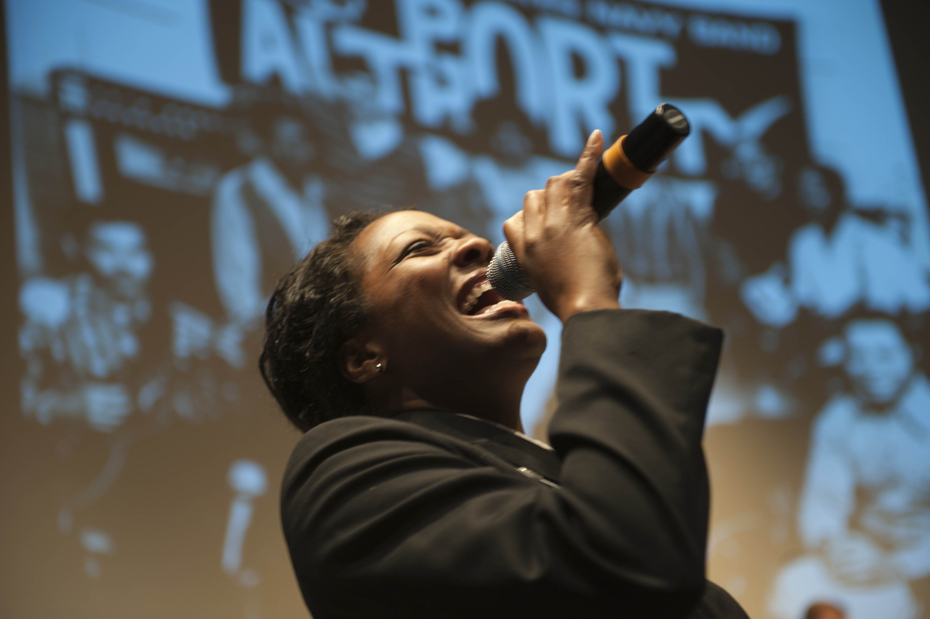 Eine Frau in einem schwarzen Kleid singt leidenschaftlich in ein Mikrofon auf der Bühne, ihre Augen sind geschlossen und ihr Gesichtsausdruck ist intensiv, während die Bühnenbeleuchtung ihr Gesicht erhellt.