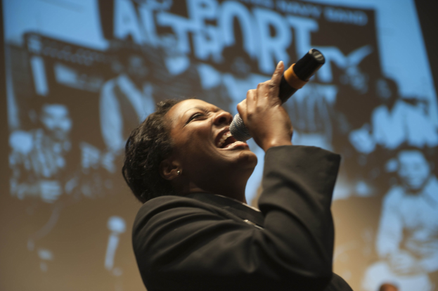 Eine Frau in einem schwarzen Kleid singt leidenschaftlich in ein Mikrofon auf der Bühne, ihre Augen sind geschlossen und ihr Gesichtsausdruck ist intensiv, während die Bühnenbeleuchtung ihr Gesicht erhellt.