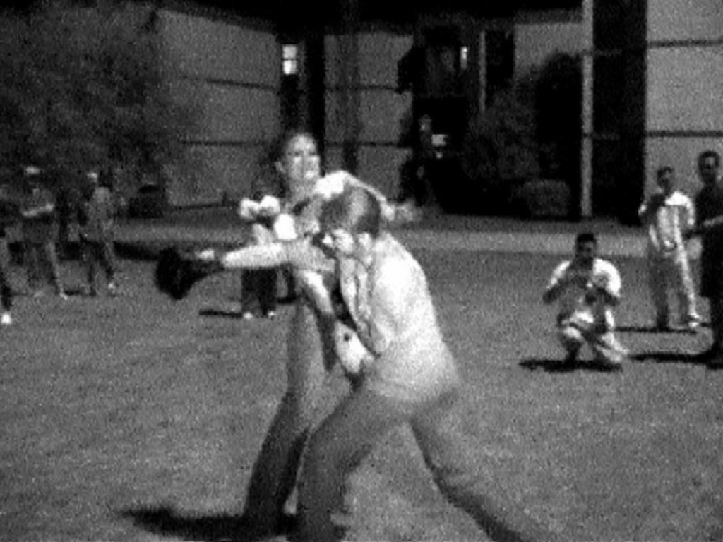 Zwei Personen boxen in der Mitte eines Schwarz-Weiß-Bildes, mit einem Gebäude und einigen Zuschauern im Hintergrund.