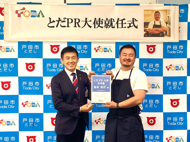 Zwei Männer mit einem Zertifikat, die lächeln, mit einem "Toda City Wins PR Award"-Banner und einem Personenfoto im Hintergrund.