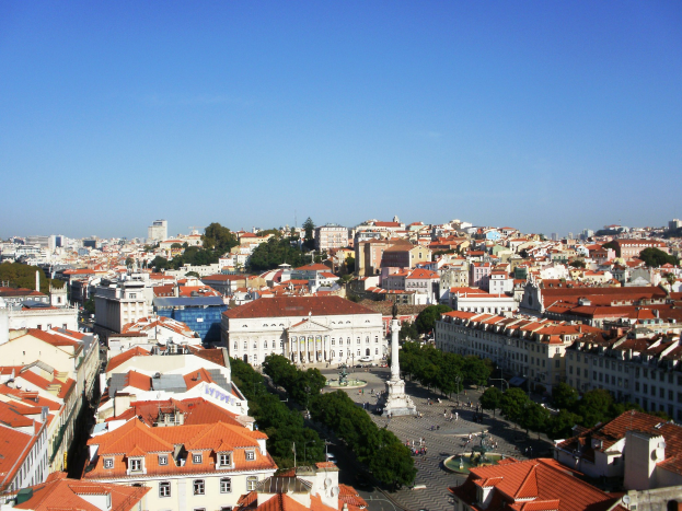 Blick auf Lissabon von einem Hügel aus, der Gebäude, Bäume, eine Statue auf einem Sockel, eine Straße mit einigen Menschen und den Himmel im Hintergrund zeigt.