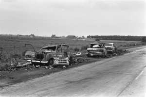Zwei Autos bei einem Zusammenstoß auf der Seite einer Straße, umgeben von Gras, Pflanzen, Bäumen und Häusern unter einem klaren Himmel.