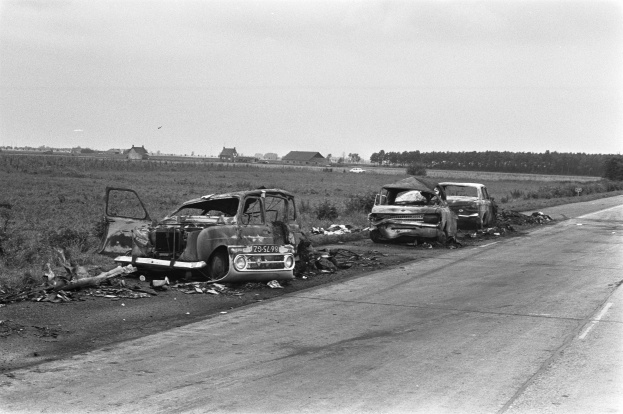 Zwei Autos bei einem Zusammenstoß auf der Seite einer Straße, umgeben von Gras, Pflanzen, Bäumen und Häusern unter einem klaren Himmel.