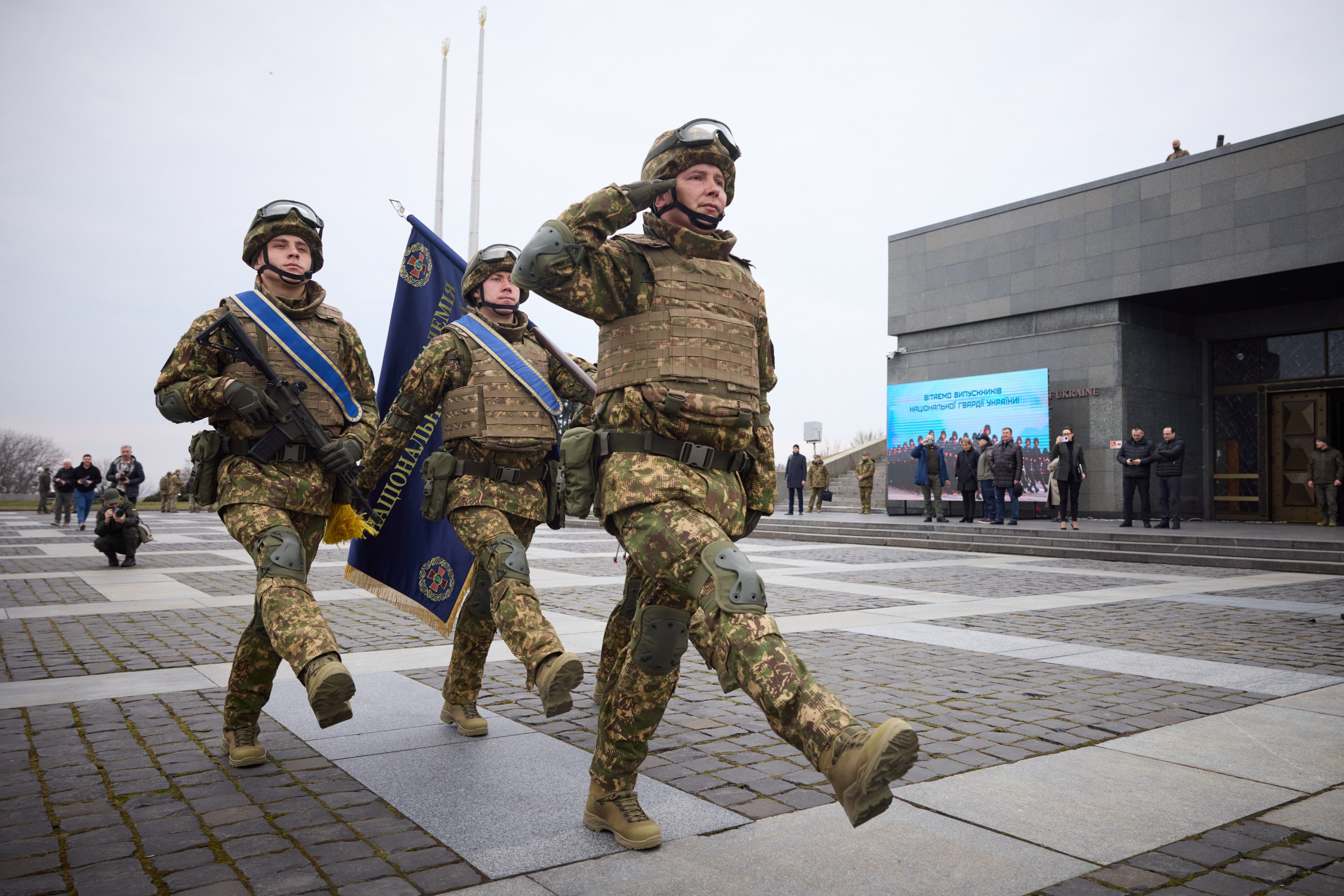 A group of Ukrainian soldiers in uniform march down a street carrying rifles and flags, with a crowd, trees, and a building in the background.