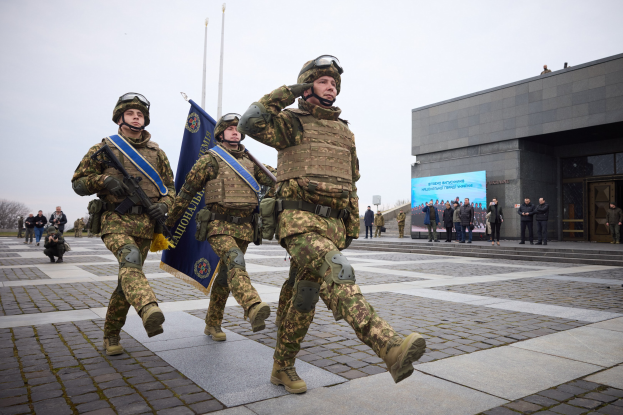 A group of Ukrainian soldiers in uniform march down a street carrying rifles and flags, with a crowd, trees, and a building in the background.