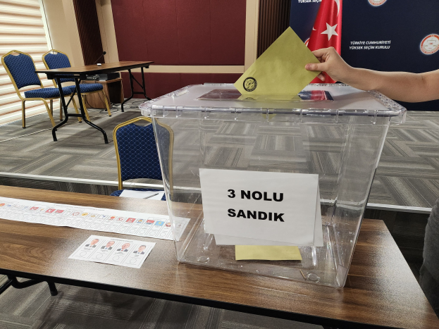 A person placing a voting paper into a ballot box on a table with chairs, a banner, and window blinds nearby.