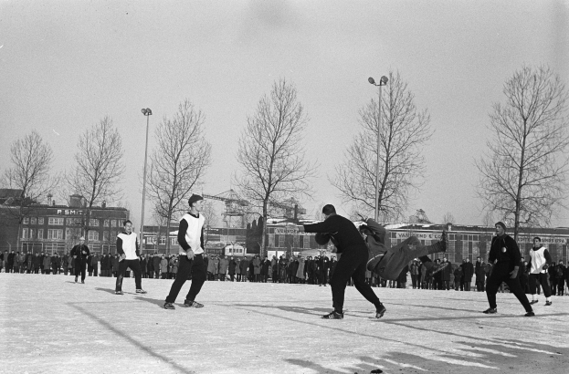 Schwarzes Bild von Menschen, die Eis-Hockey im Schnee spielen, mit Bäumen, Gebäuden, Fenstern, Laternenmasten und einem klaren Himmel im Hintergrund.