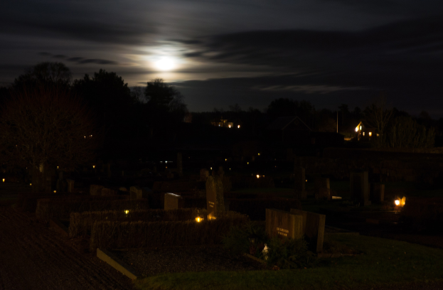Friedhof bei Nacht mit Vollmond am Himmel, Gräber und Bäume durch künstliche Lichter beleuchtet.