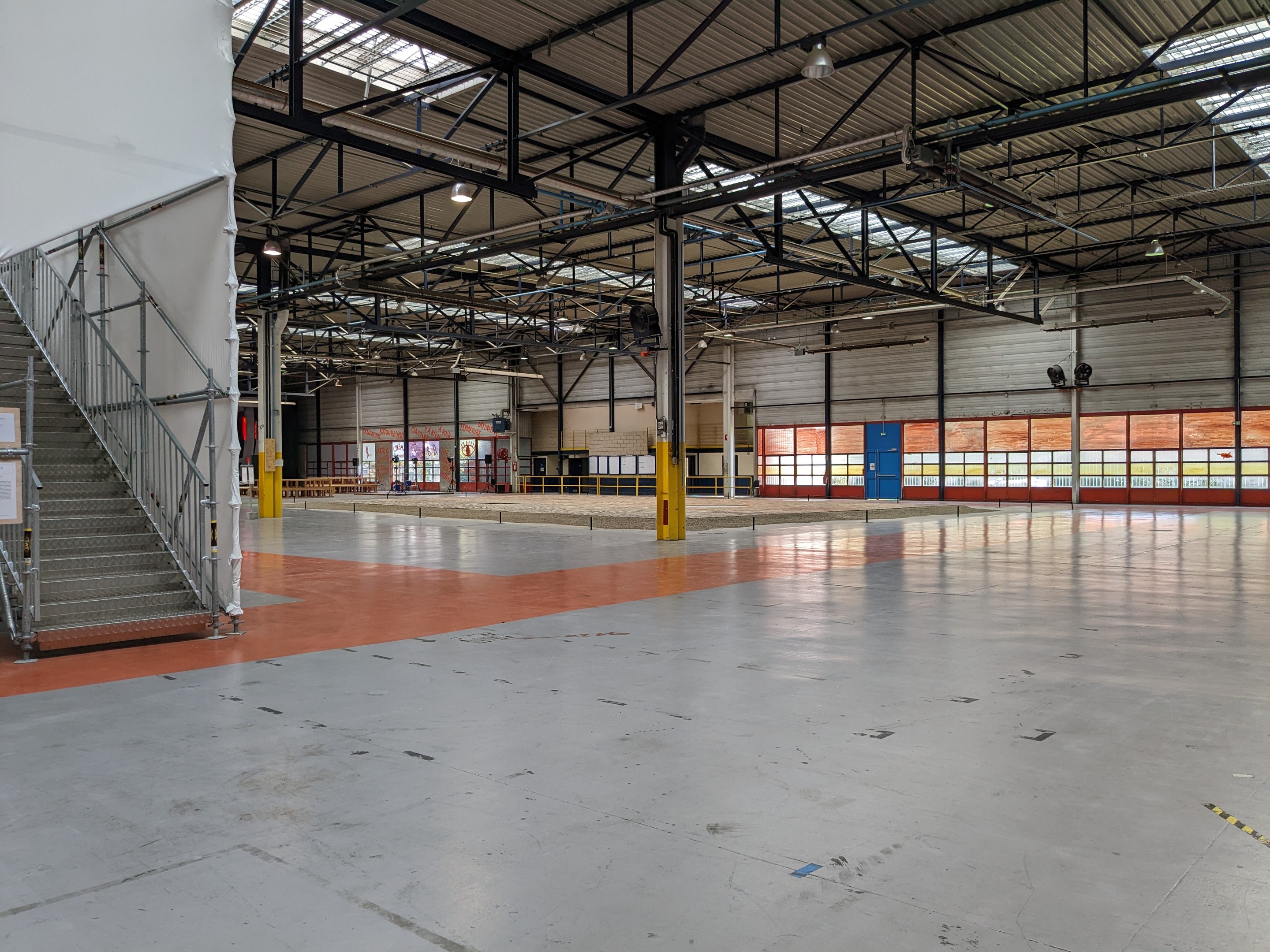 Industrielles Lager-Innenraum mit großen Fenstern, Metall-Stützpfosten, einer Treppe mit Geländer, einem Schild, Deckenlampen und zusätzlichen Pfosten.