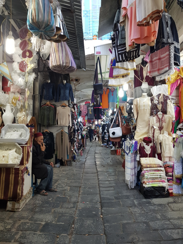 A bustling street market in Jerusalem's old city with stalls selling clothes, bags, lights, and other items, and people browsing the selection.
