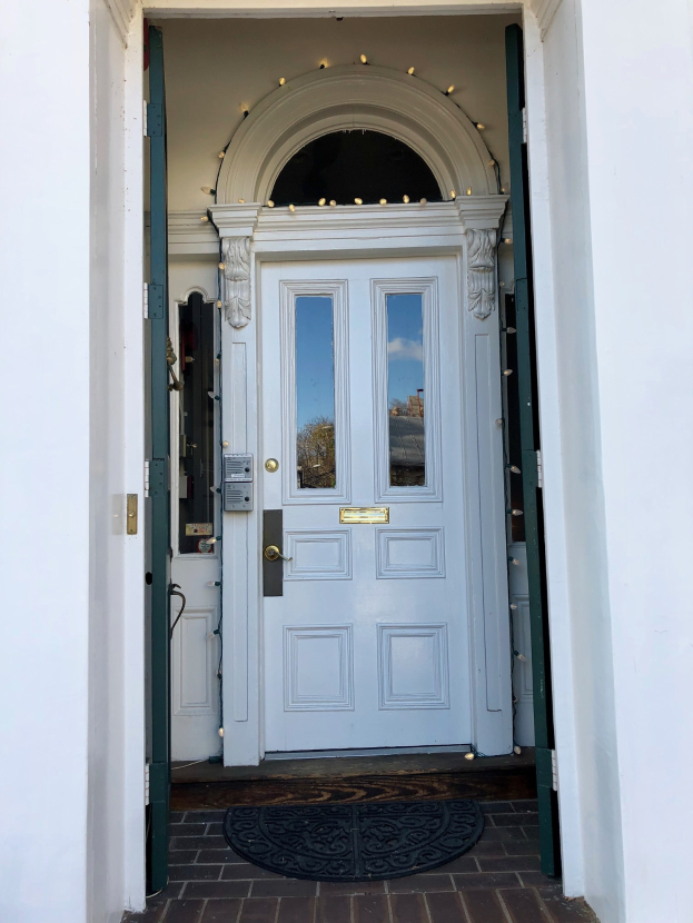 Ein weißes Haus mit einer blauen Haustür, einem Türvorleger und dekorativen Lichtern an der Tür.