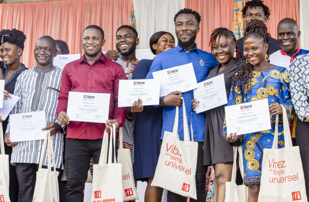 A group of smiling people holding certificates and standing with bags near curtains and a wall, celebrating an award win.