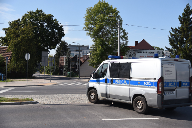 Polizeifahrzeug auf grasbewachsener Straße mit Hinweistafeln, Laternenmasten, Bäumen, Gebäuden und bewölktem Himmel im Hintergrund.