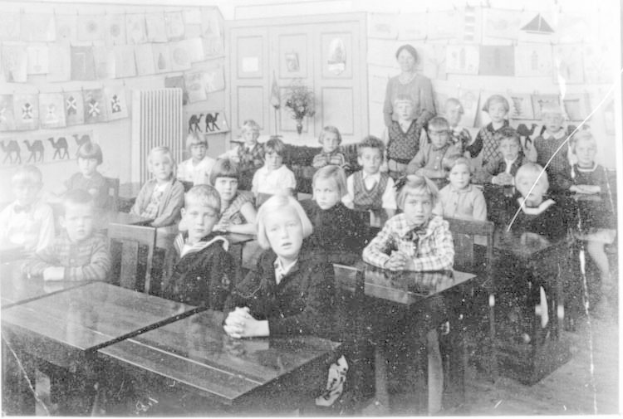 Schwarzes und weißes Foto einer Klasse mit Kindern auf Bänken sitzend und auf dem Boden stehend, mit einem Blumenstrauß, Papieren an einer Tafel, einer Tür und Deckenlampen an der Rückwand.
