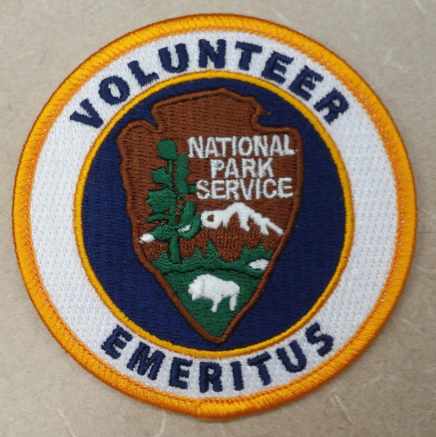 Ein rundes Patch mit einem blauen Rand und einem weißen Hintergrund, das eine zentrierte schwarze Schrift "Volunteer National Park Service Emeritus" zeigt, umgeben von einem gelben Kreis.