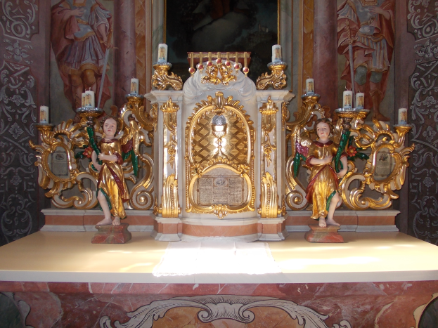 Ein Altar in einer Kirche mit Skulpturen von Menschen und Kerzen auf Ständern, mit einem Bild an der Wand dahinter.