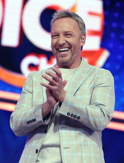A man in a suit clapping on a stage with "The Voice" logo in the background.