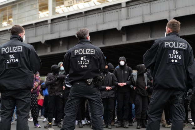 Polizisten in schwarzer Uniform und Masken stehen vor einer Menge während einer Demonstration, im Hintergrund ist eine Brücke und ein Gebäude zu sehen.