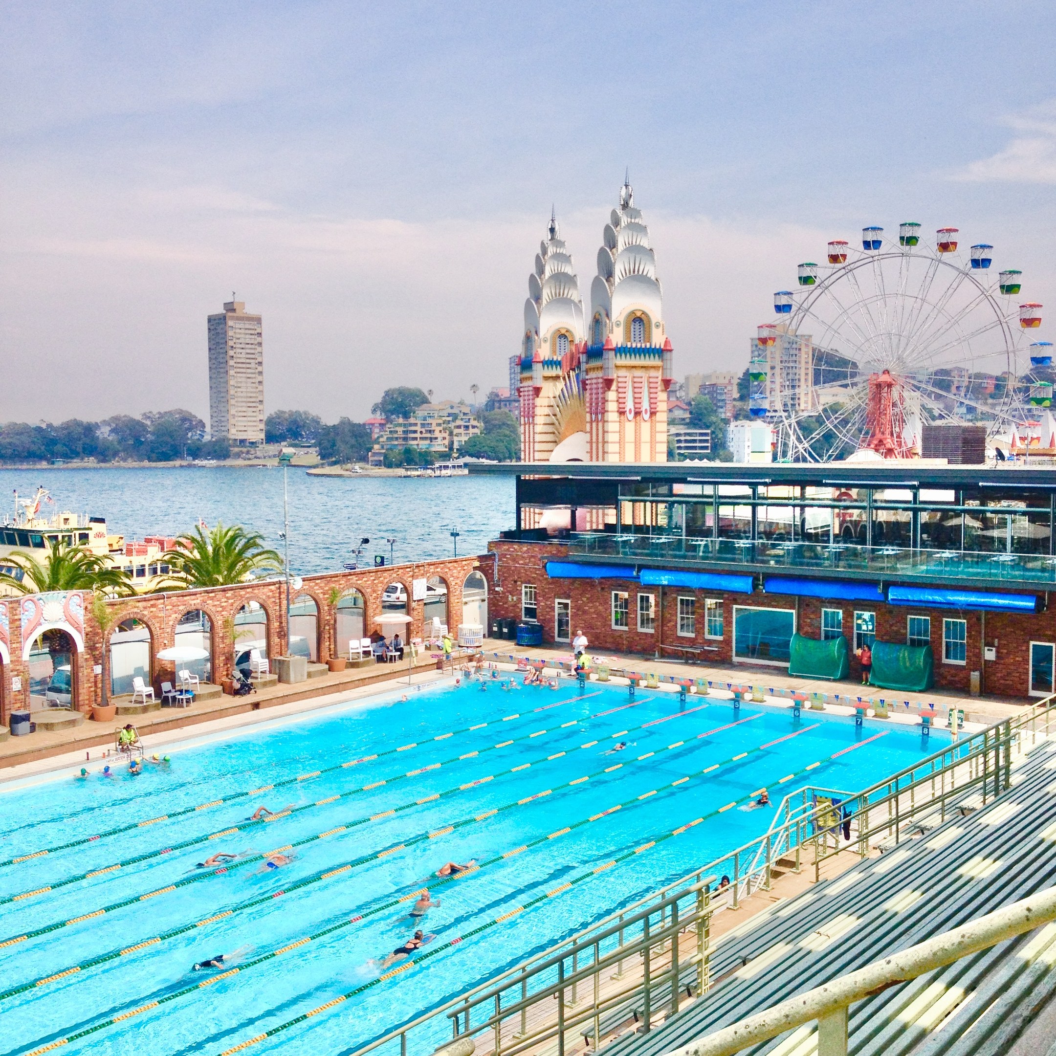 Large swimming pool with people swimming, surrounded by railings and benches, with a giant ferris wheel, buildings, trees, and a cloudy sky in the background.