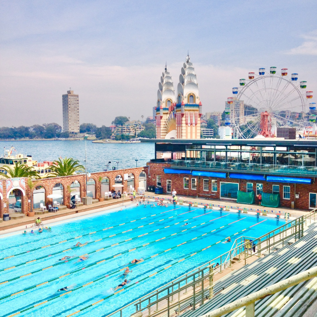 Large swimming pool with people swimming, surrounded by railings and benches, with a giant ferris wheel, buildings, trees, and a cloudy sky in the background.