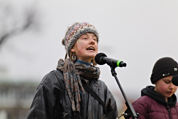Eine Frau in schwarzer Jacke, Schal und Mütze singt in ein Mikrofon auf der Bühne, neben ihr rechts eine Person in einer maronenfarbenen Jacke und schwarzer Mütze, im Hintergrund eine unscharfe Menge.