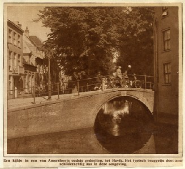 Altes Schwarz-Weiß-Foto einer Brücke, die einen Kanal überspannt, mit Radfahrern, einem Baum im Hintergrund, Gebäuden auf beiden Seiten und Text am unteren Rand.