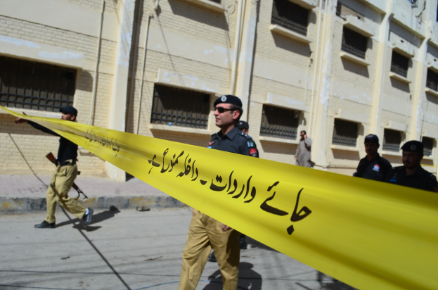 Gruppe von Polizisten in Uniform vor einem Gebäude mit gelbem Band, einer Person mit Waffe, mit sichtbarem Himmel und Gebäudedetails im Hintergrund.
