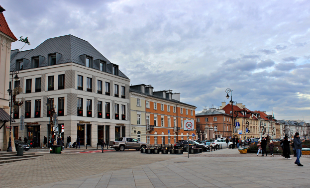 Ein belebter Stadtplatz mit mehreren Gebäuden, Fahrzeugen, Fußgängern, Straßenmöblierung und Bäumen unter einem bewölkten Himmel.