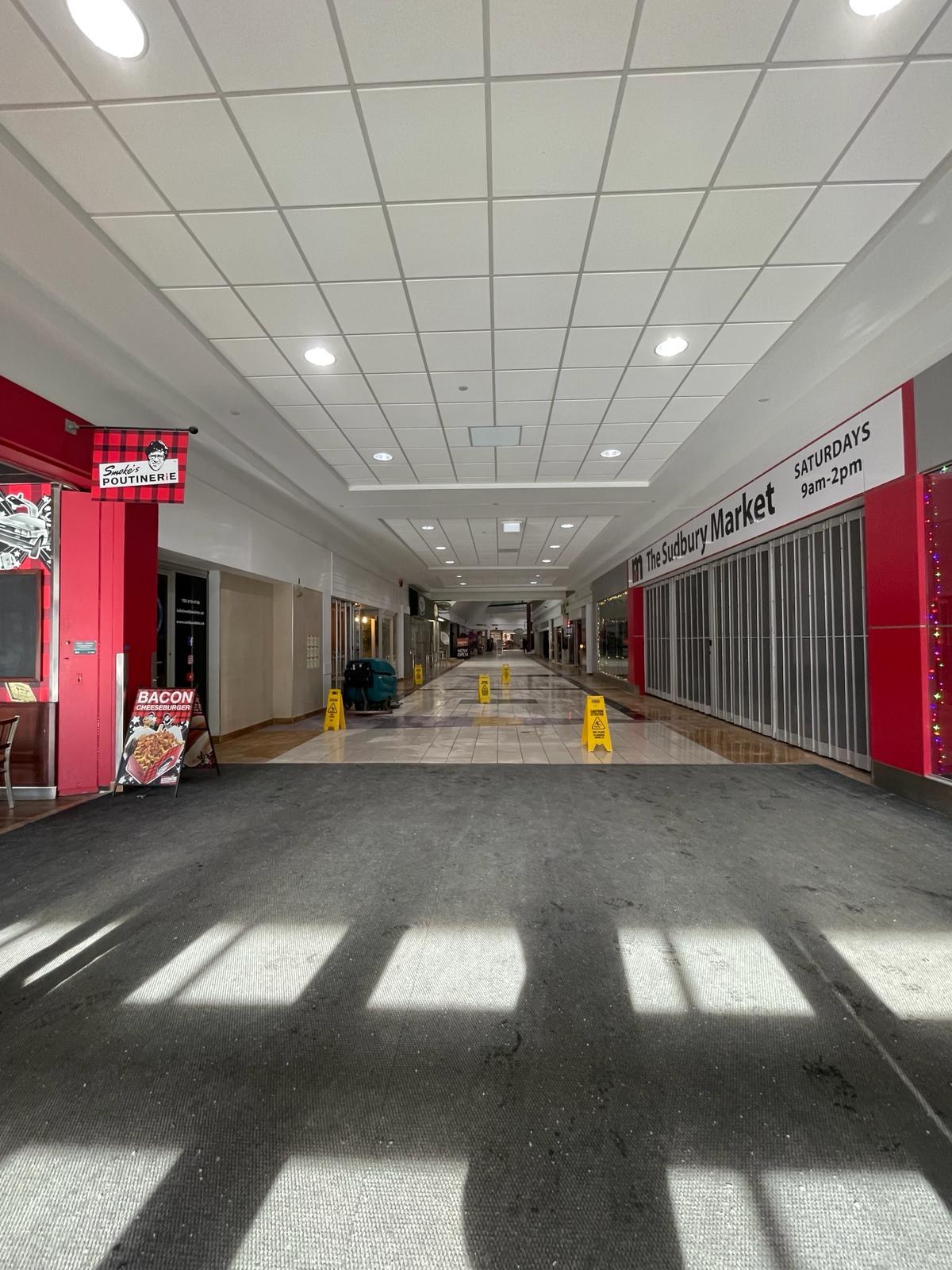 Innenraum eines Einkaufszentrums mit Deckenbeleuchtung, einem Stuhl und einer Tafel mit der Aufschrift "Sydbury Market - Vorher und Nachher" links und verstreuten Gegenständen rechts und auf dem Boden.