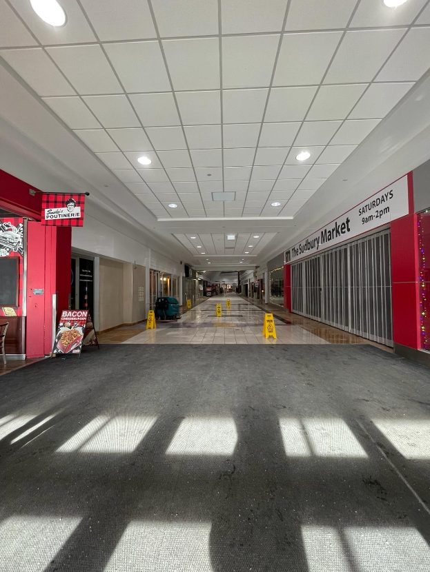 Innenraum eines Einkaufszentrums mit Deckenbeleuchtung, einem Stuhl und einer Tafel mit der Aufschrift "Sydbury Market - Vorher und Nachher" links und verstreuten Gegenständen rechts und auf dem Boden.