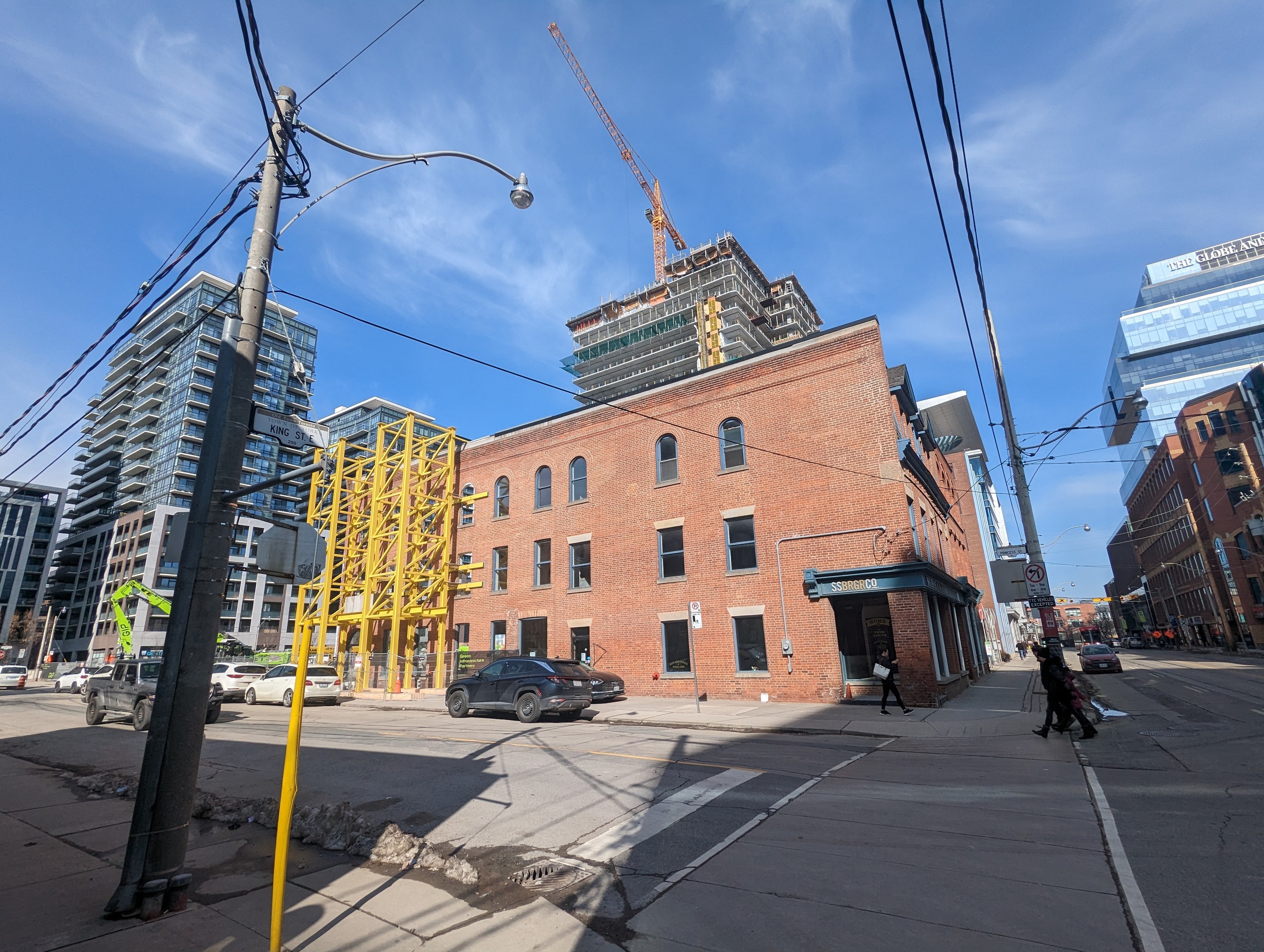 Eine belebte Stadtstraße mit fahrenden Autos, Fußgängern auf dem Gehweg, Strommasten, Straßenlaternen und Gebäuden, mit einem Baukran gegen einen bewölkten Himmel.