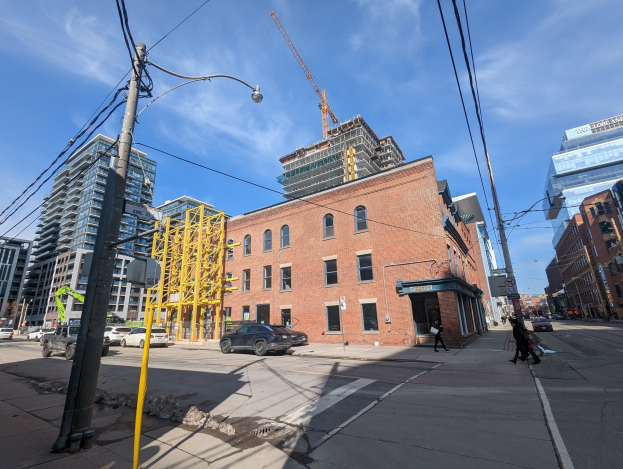 Eine belebte Stadtstraße mit fahrenden Autos, Fußgängern auf dem Gehweg, Strommasten, Straßenlaternen und Gebäuden, mit einem Baukran gegen einen bewölkten Himmel.