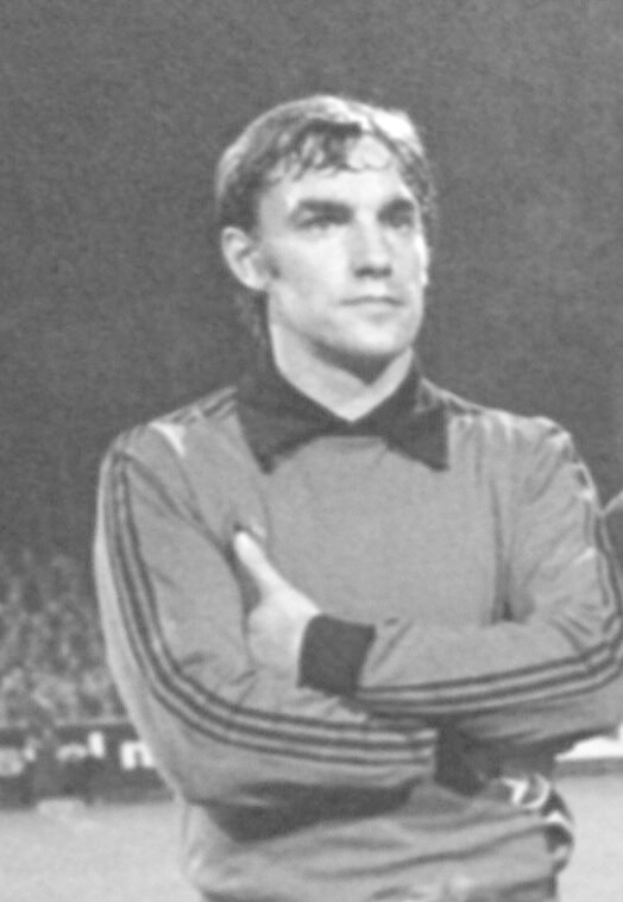Black and white photo of a man standing on a soccer field with arms crossed, surrounded by seated spectators.