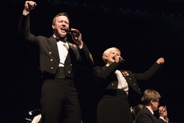 Zwei Militärangehörige in Uniform singen auf der Bühne, wobei einer ein Mikrofon hält und ein anderer einen schwarzen Blazer und ein weißes Hemd trägt, während andere im Hintergrund sitzen.