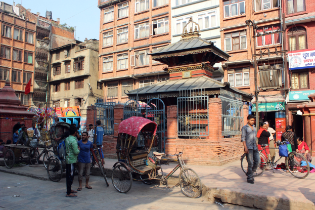 A bustling street in Kathmandu, Nepal, with pedestrians, rickshaws, bicycles, and vehicles alongside tall buildings, a gate, a flag, and a clear blue sky.