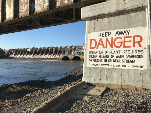Eine Brücke mit einem "Achtung"-Schild, umgeben von Wasser, mit Gebäuden und einem klaren blauen Himmel im Hintergrund.