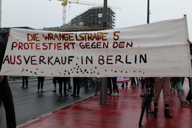 Group of people holding a banner reading "Die Wrangellstraße 5 Protestiert Gegen den Ausverkauf in Berlin" on a city street with bicycles, poles, buildings, a crane, and sky in the background.