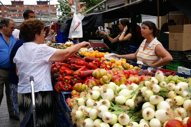 Gemüse in Plastikbehältern, eine Gruppe von Menschen, Gebäude, Bäume, eine Kartonbox und der Himmel.