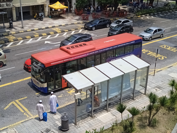 Ein roter und blauer Bus hält an einer Bushaltestelle auf einer Straße mit Fahrzeugen, Fußgängern, einem Schuppen, Pflanzen, Bäumen, Gebäuden und verschiedenen Gegenständen im Hintergrund.
