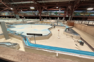 Ein großes Indoor-Wasserpark mit mehreren Wasserrutschen, Pfosten, Beschilderung, Deckenleuchten und Wandgemälde, das als Minigolfplatz konzipiert ist.