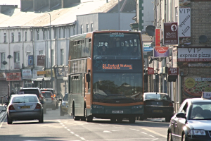 Eine Straße mit Autos und einem Bus vor Gebäuden mit Wänden, Fenstern, Tellern und Dächern, die Plakate und Banner an den Wänden und einen Pfahl mit einer Straßenlaterne zeigen.