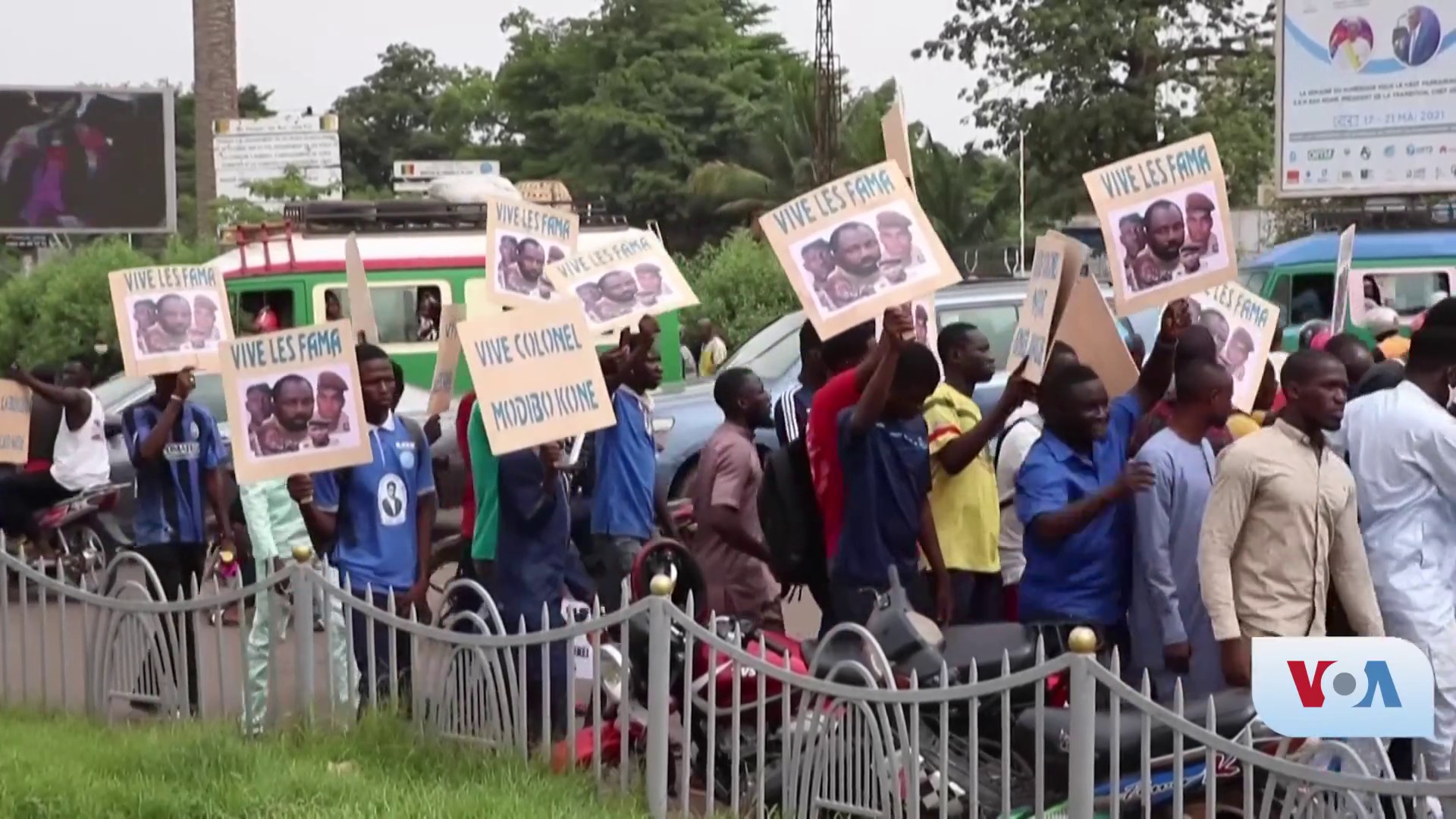 Eine Gruppe von Menschen h├Ąlt Schilder, wahrscheinlich protestierend, vor einem Zaun mit Fahrzeugen, B├Ąumen, Pf├Âhlen, Absperrungen und einem Bildschirm im Hintergrund, unter einem sichtbaren Himmel mit Gras am unteren Bildrand.